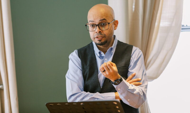 A bald man in a dress shirt and vest, and with glasses, stands at a podium delivering a sermon.
