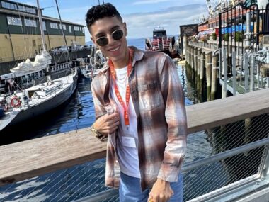 A stylish young man stands on a boardwalk with boats and water behind him, wearing dark, round sunglasses and a flannel shirt.