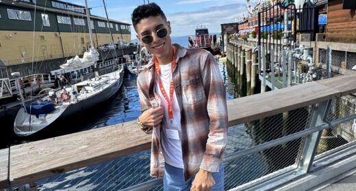 A stylish young man stands on a boardwalk with boats and water behind him, wearing dark, round sunglasses and a flannel shirt.