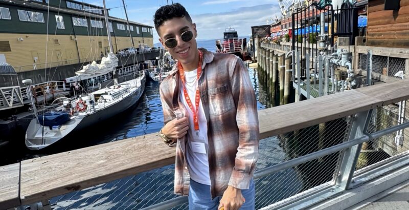 A stylish young man stands on a boardwalk with boats and water behind him, wearing dark, round sunglasses and a flannel shirt.