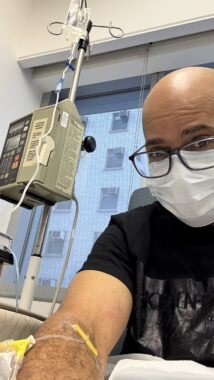 A man in a hospital takes a selfie while receiving an infusion. He's wearing glasses and a mask. 