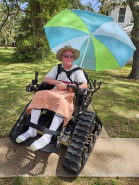 A man is strapped into an all-terrain wheelchair and sitting in the shade of large trees on a sunny day. He also has a green and blue umbrella affixed to the back of his chair to provide additional shade, and he's wearing socks and is covered in a light blanket.