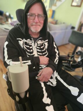 A man with glasses and sitting in a wheelchair in his living room stares intensely at the camera, which could be intimidating or off-putting if not for the fact that he's wearing a full-body skeleton costume for Halloween.