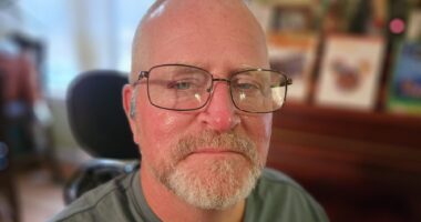 A close-up cropped shot of a middle-aged man with glasses staring at the camera. Blurry background images like picture frames indicate that it's the inside of someone's home.