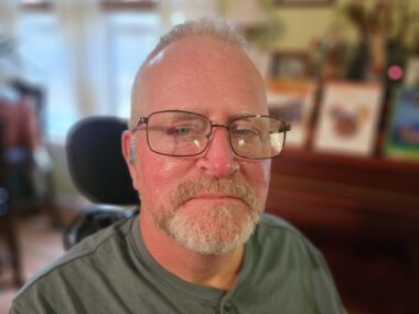 A close-up cropped shot of a middle-aged man with glasses staring at the camera. Blurry background images like picture frames indicate that it's the inside of someone's home.