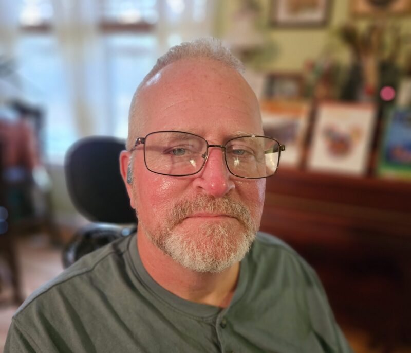 A close-up cropped shot of a middle-aged man with glasses staring at the camera. Blurry background images like picture frames indicate that it's the inside of someone's home.