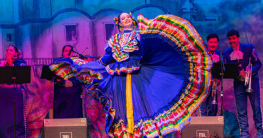 A woman wearing a traditional Mexican dress performs a dance onstage.