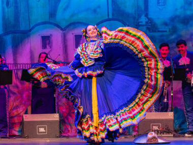 A woman wearing a traditional Mexican dress performs a dance onstage.