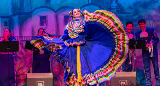 A woman wearing a traditional Mexican dress performs a dance onstage.