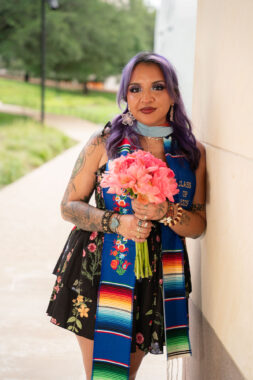 A woman leans against a wall while holding a bouquet of pink flowers. She's wearing a black sundress and a sash to celebrate earning her master's degree.
