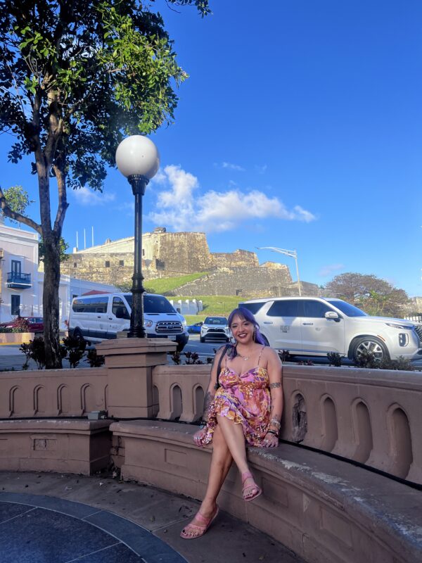 A woman sits on a bench outside next to a busy street in Puerto Rico.