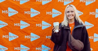 A young woman poses in front of an orange banner covered with the MS Walk logo. She's wearing a black jacket, a crossbody bag, and is holding a Starbucks cup in her right hand.