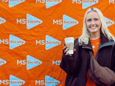 A young woman poses in front of an orange banner covered with the MS Walk logo. She's wearing a black jacket, a crossbody bag, and is holding a Starbucks cup in her right hand.