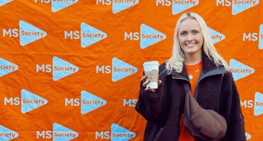 A young woman poses in front of an orange banner covered with the MS Walk logo. She's wearing a black jacket, a crossbody bag, and is holding a Starbucks cup in her right hand.