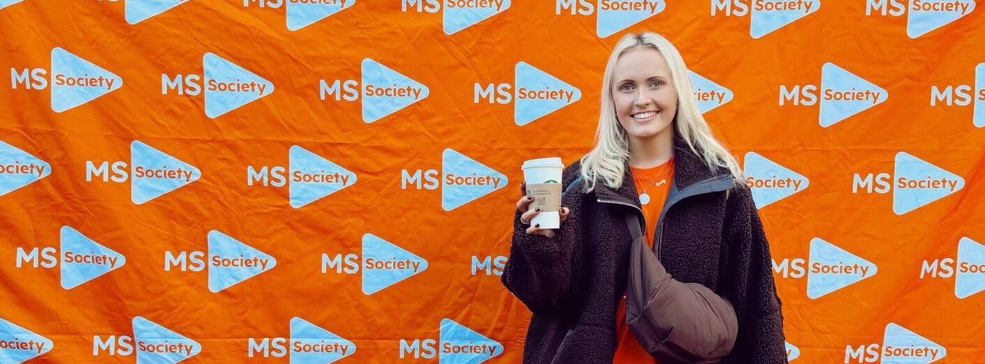 A young woman poses in front of an orange banner covered with the MS Walk logo. She's wearing a black jacket, a crossbody bag, and is holding a Starbucks cup in her right hand.