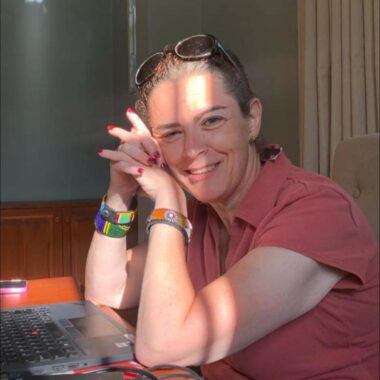 A woman sits at a desk with her head gently resting on folded hands, propped up by her elbows. Sunlight shining in through a nearby window casts an interesting contrast of light and shadow on her face. 