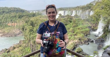 A woman with a professional camera stands on a steel railing overlooking a beautiful, vast, river landscape with numerous large waterfalls and green vegetation. She's smiling and wears an expression of elated awe.
