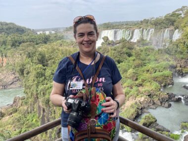 A woman with a professional camera stands on a steel railing overlooking a beautiful, vast, river landscape with numerous large waterfalls and green vegetation. She's smiling and wears an expression of elated awe.