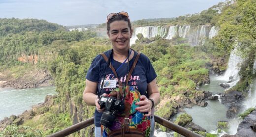 A woman with a professional camera stands on a steel railing overlooking a beautiful, vast, river landscape with numerous large waterfalls and green vegetation. She's smiling and wears an expression of elated awe.
