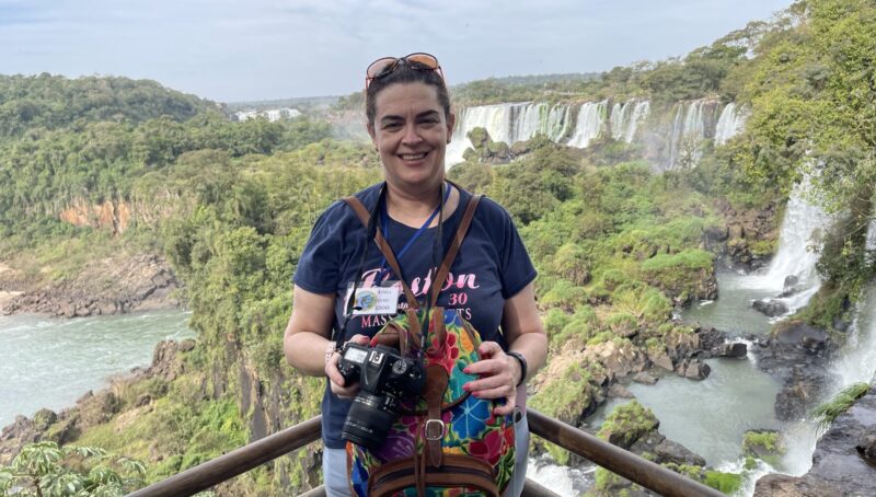 A woman with a professional camera stands on a steel railing overlooking a beautiful, vast, river landscape with numerous large waterfalls and green vegetation. She's smiling and wears an expression of elated awe.