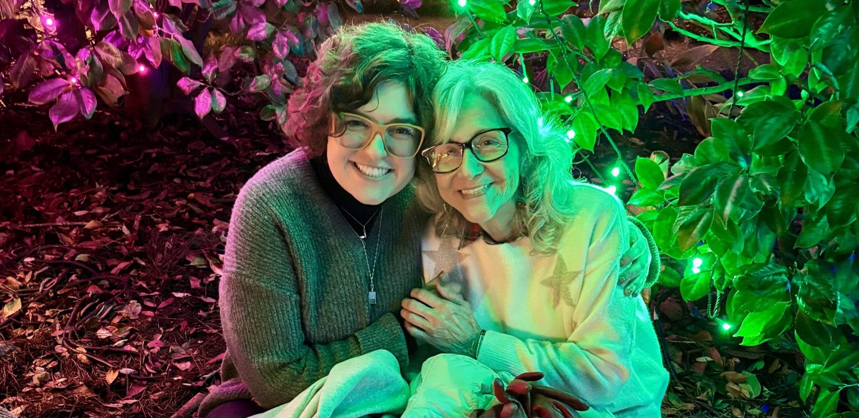 A woman and her grandmother embrace each other in a garden with some trippy pinkish-red and green lights casting a dynamic glow over the two's faces.