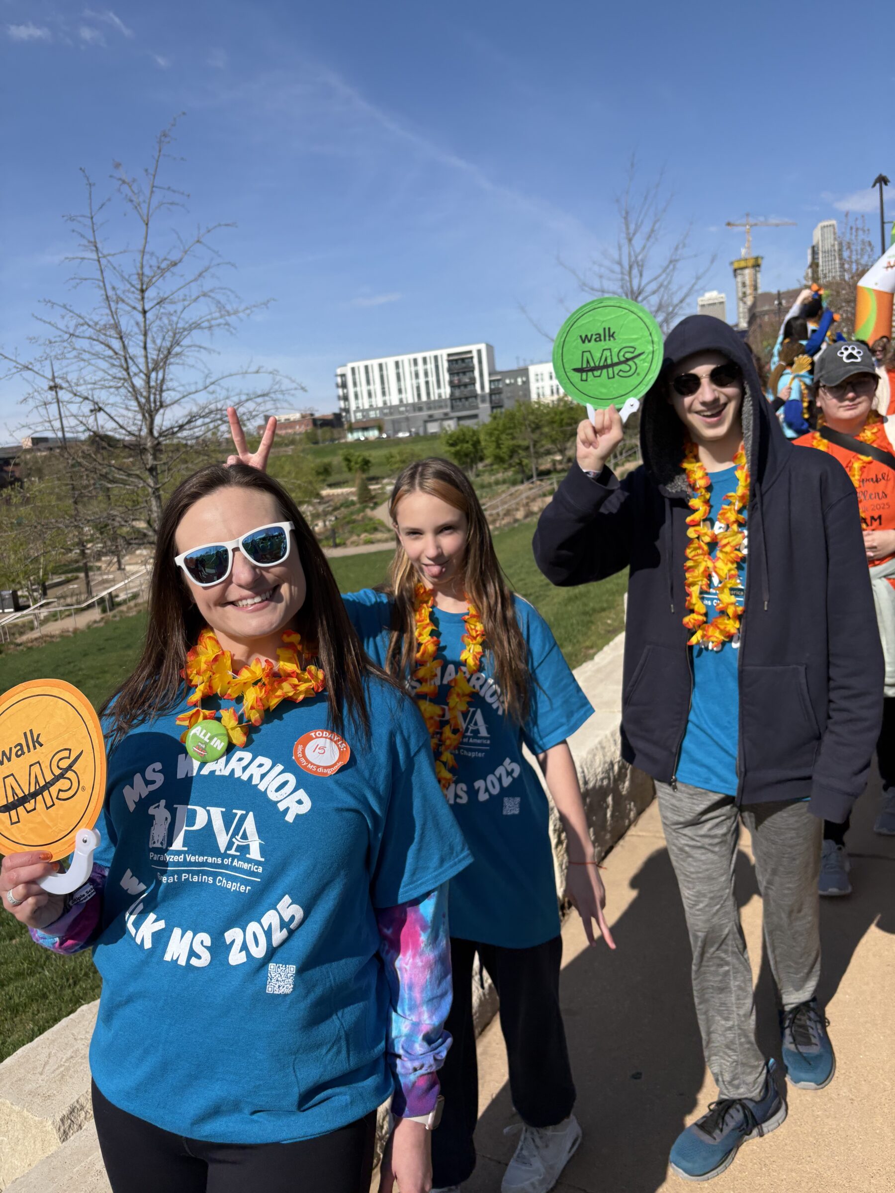 A mother, young daughter and young son, pose on a sunny day during an advocacy event. They're all wearing blue MS advocacy T-shirts and colorful lei.