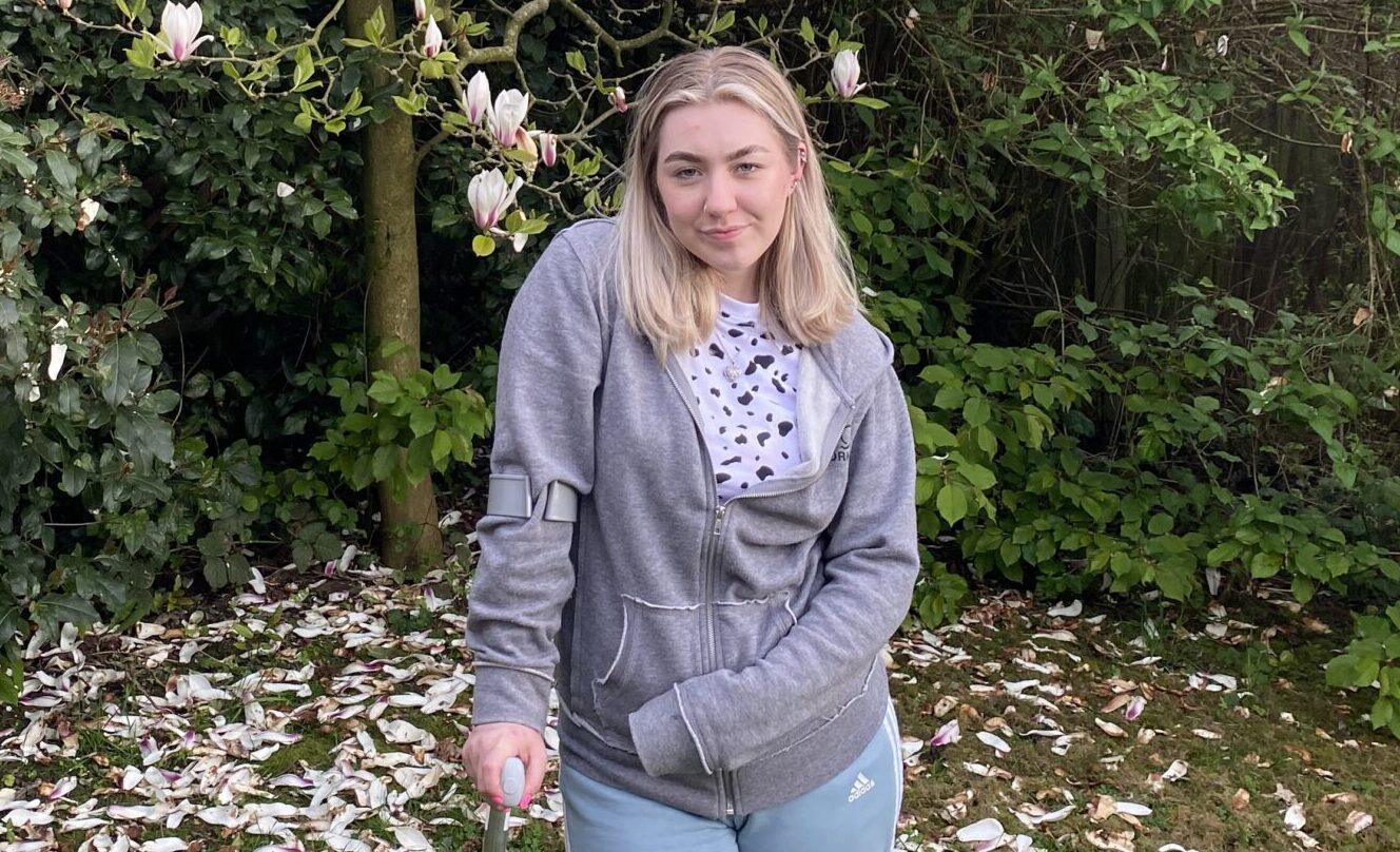A young woman with blond hair is pictured in her garden, leaning on a cane.