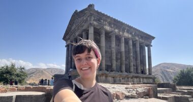 A woman takes a selfie in front of an ancient unidentified structure outdoors, probably somewhere in Europe, likely Greece.