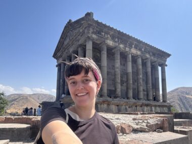 A woman takes a selfie in front of an ancient unidentified structure outdoors, probably somewhere in Europe, likely Greece.