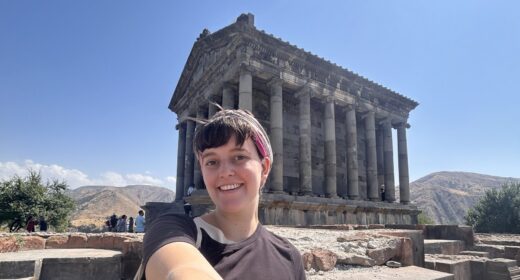 A woman takes a selfie in front of an ancient unidentified structure outdoors, probably somewhere in Europe, likely Greece.