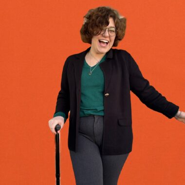 A woman poses in a lively and energetic manner in a photo that's photoshopped with a bright orange background. She swings her left arm back in a playful manner, and holds a cane in her right. 