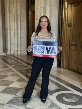 A woman stands in what appears to be the halls of Congress holding a sign that says "We Choose VA." 