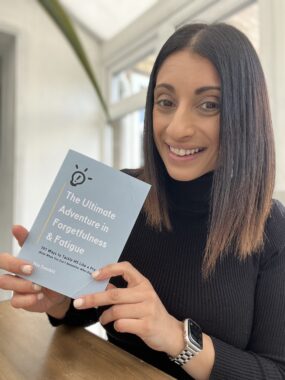 A smiling woman sits at a table and holds a copy of a gray paperback book with white lettering. 