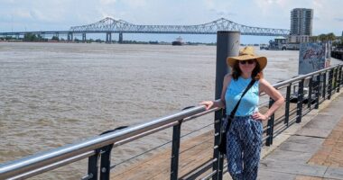 A woman stands on a walkway next to a large body of water, possibly a river. She has one hand on a railing, and her other hand rests on her hip. There's a large bridge in the background.