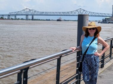 A woman stands on a walkway next to a large body of water, possibly a river. She has one hand on a railing, and her other hand rests on her hip. There's a large bridge in the background.