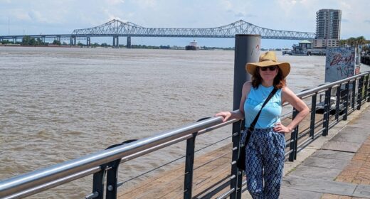 A woman stands on a walkway next to a large body of water, possibly a river. She has one hand on a railing, and her other hand rests on her hip. There's a large bridge in the background.