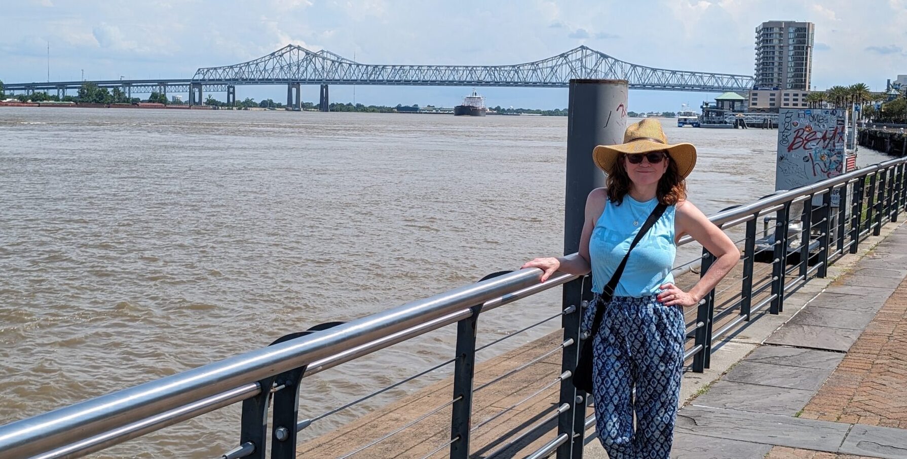 A woman stands on a walkway next to a large body of water, possibly a river. She has one hand on a railing, and her other hand rests on her hip. There's a large bridge in the background.
