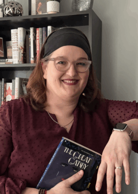 A woman with a black headband and stylish glasses smiles as she leans on a shelf while holding a copy of the book "The Great Gatsby." Directly behind her is a bookshelf.