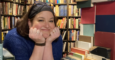 A really cool photo of a woman peering in between stacks and stacks of books, designed almost like a window of a house. She rests her chin on her two hands and flashes a warm, friendly smile, her glasses perched on the top of her head.