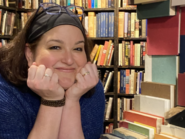 A really cool photo of a woman peering in between stacks and stacks of books, designed almost like a window of a house. She rests her chin on her two hands and flashes a warm, friendly smile, her glasses perched on the top of her head.