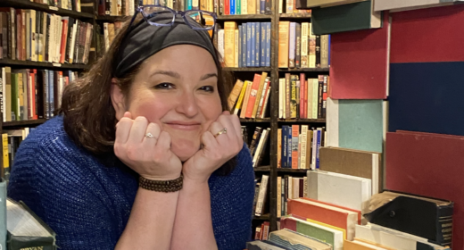 A really cool photo of a woman peering in between stacks and stacks of books, designed almost like a window of a house. She rests her chin on her two hands and flashes a warm, friendly smile, her glasses perched on the top of her head.