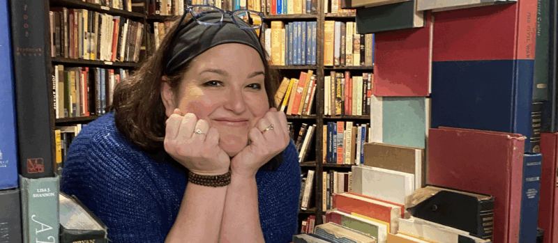 A really cool photo of a woman peering in between stacks and stacks of books, designed almost like a window of a house. She rests her chin on her two hands and flashes a warm, friendly smile, her glasses perched on the top of her head.