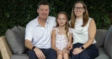 A family sits for a tightly framed family photo with a man sitting next to a woman, and a young girl sitting on one knee of each parent in the middle.