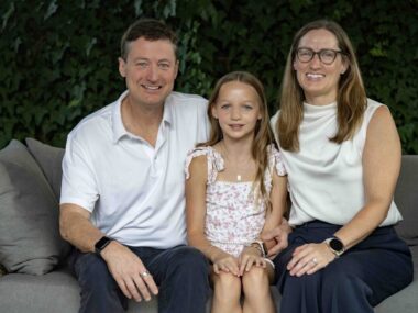 A family sits for a tightly framed family photo with a man sitting next to a woman, and a young girl sitting on one knee of each parent in the middle.