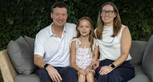 A family sits for a tightly framed family photo with a man sitting next to a woman, and a young girl sitting on one knee of each parent in the middle.