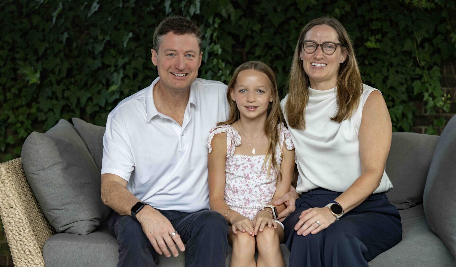 A family sits for a tightly framed family photo with a man sitting next to a woman, and a young girl sitting on one knee of each parent in the middle.