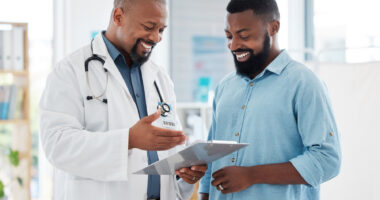 A doctor gestures at a clipboard while showing a patient his records.