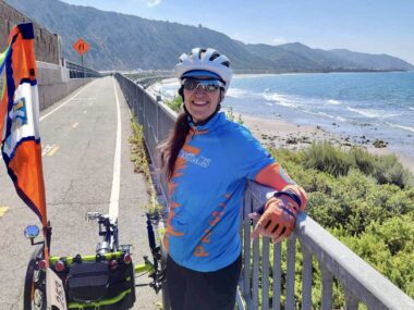 A woman in cycling gear - including cycling clothing, a bike helmet, and sunglasses - pauses to lean on a railing along the side of a road that runs parallel to the ocean. Her bike is parked next to her, and the woman is smiling broadly. It's a sunny day and the ocean waters look inviting. On the horizon, mountains rise from the coast.