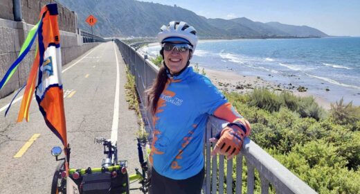 A woman in cycling gear - including cycling clothing, a bike helmet, and sunglasses - pauses to lean on a railing along the side of a road that runs parallel to the ocean. Her bike is parked next to her, and the woman is smiling broadly. It's a sunny day and the ocean waters look inviting. On the horizon, mountains rise from the coast.
