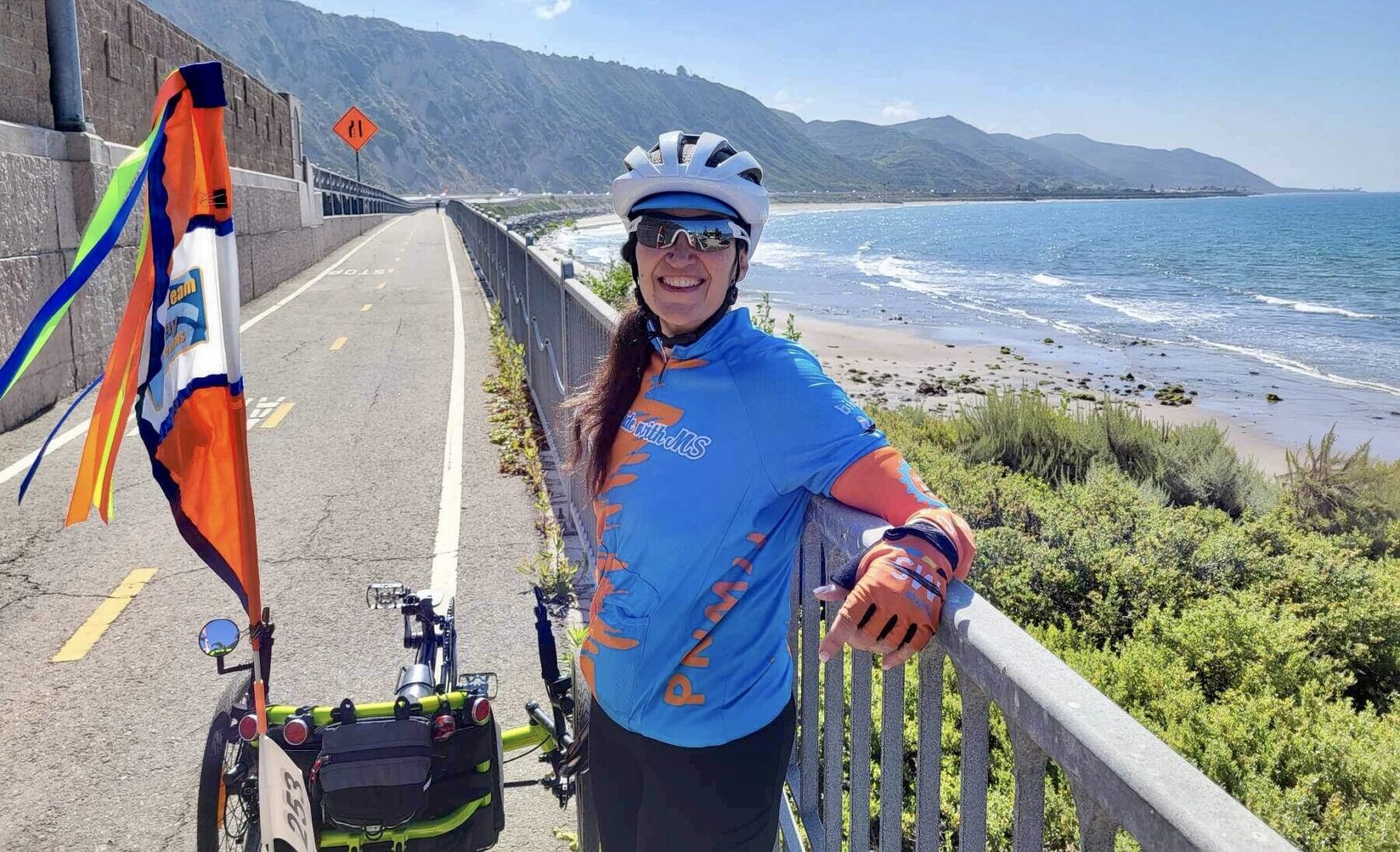 A woman in cycling gear - including cycling clothing, a bike helmet, and sunglasses - pauses to lean on a railing along the side of a road that runs parallel to the ocean. Her bike is parked next to her, and the woman is smiling broadly. It's a sunny day and the ocean waters look inviting. On the horizon, mountains rise from the coast.